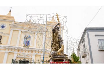 Il santo patrono, in primo piano, è raffigurato in una statua che lo ritrae mentre tiene una palma nella mano destra, simbolo di vittoria e martirio. Alle sue spalle si erge la Basilica, la chiesa madre del patrono, con la sua facciata imponente che domina la scena. Sullo sfondo, le luminarie che decorano la piazza creano una suggestiva atmosfera di festa, rendendo ancora più luminosa e vivace la celebrazione patronale.