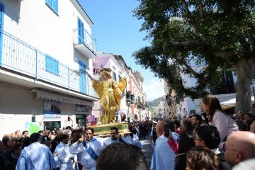 La foto mostra un momento della tradizionale Corsa dell'Angelo a Forio, con un angelo in volo, circondato da una folla entusiasta che celebra l'evento religioso e culturale.