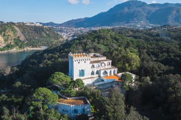 Villa La Colombaia, immersa nel verde del Bosco di Zaro, si distingue per il suo fascino e stile che si fondono armoniosamente con la bellezza naturale che la circonda. Alle sue spalle si estende il mare cristallino della Baia di San Montano, mentre un tratto dell’isola d’Ischia e le sue montagne completano il paesaggio. Il cielo azzurro e limpido di una splendida giornata esalta la serenità e la magnificenza del luogo.